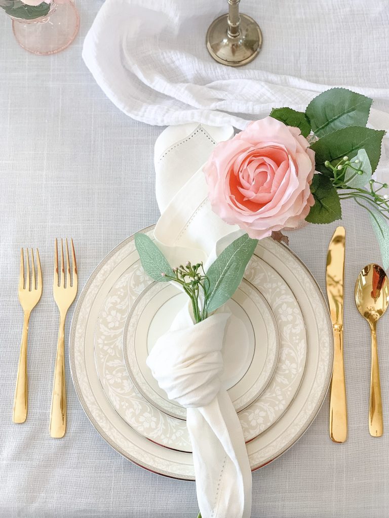An elegant place setting with ivory Noritake china, gold flatware, ivory linen napkins, and small pink rose arrangements.