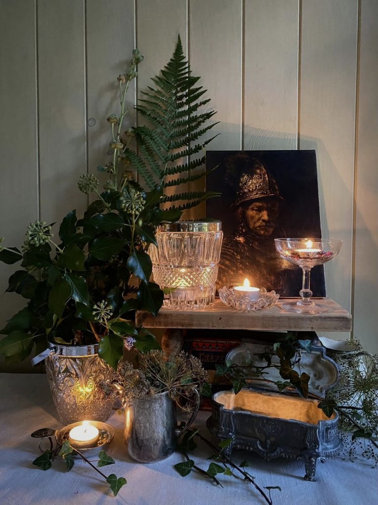 Various Crystal Ice Buckets and other glassware are filled with candles on a counter and shelf and styled with greenery and a moody portrait.