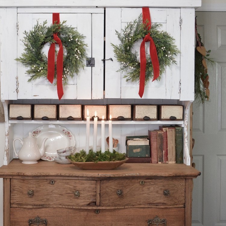 A primitive white upper cabinet is decorated with 2 Christmas wreath