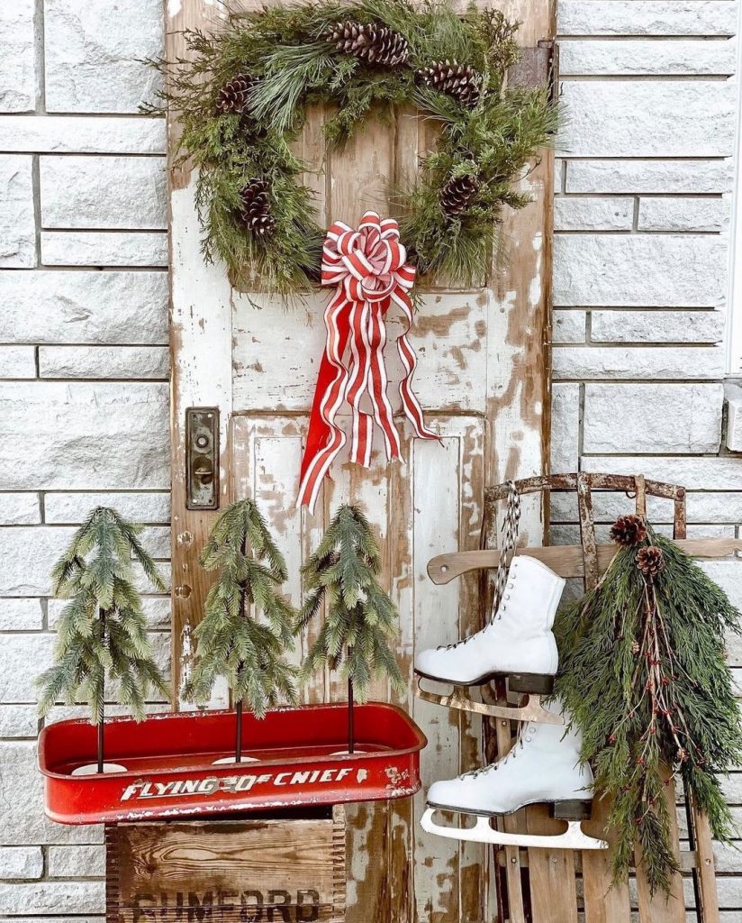A weathered vintage door holds a natural evergreen wreath and sits behind a red wagon filled with 3 small Christmas trees. A vintage sled holding ice skates leans on the door.
