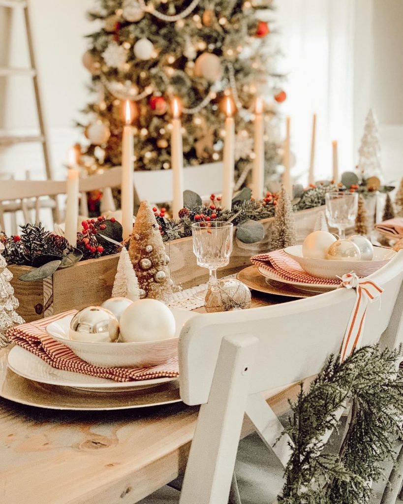An angled view of two placesettings and a Christmas centerpiece filled with greenery and lit candles. A white dining chair is in front with a wreath tied with red and white ribbon.