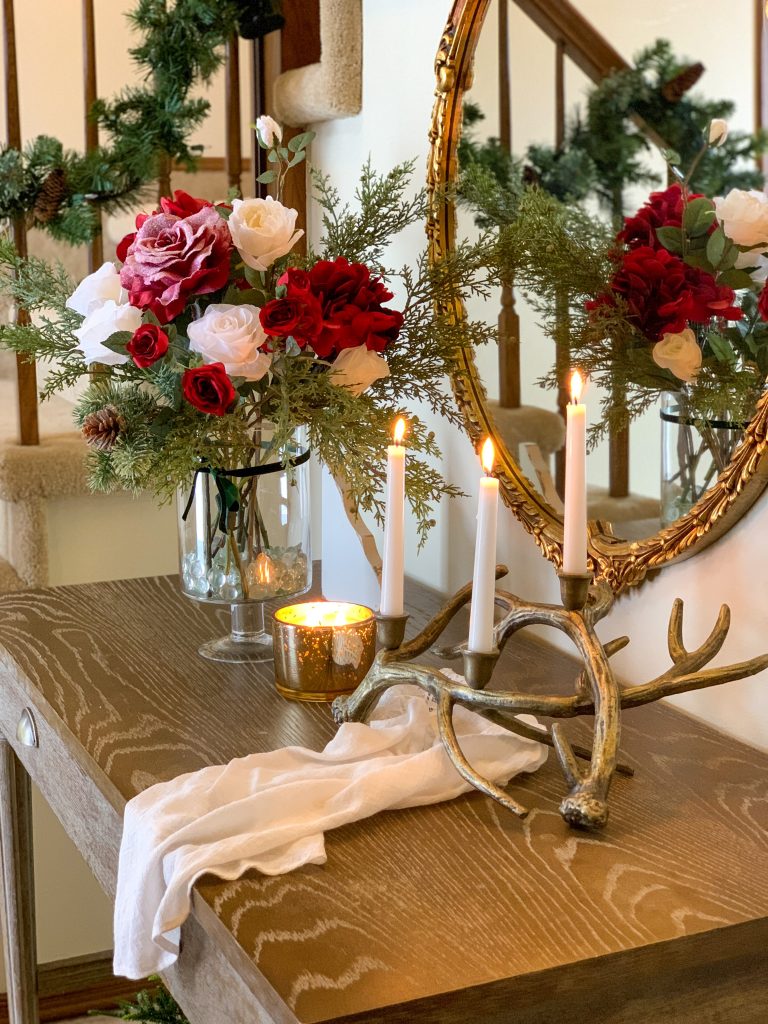 A corner view of a entry table with a Christmas arrangement, brass antler candelabra and lit candle rest on top.