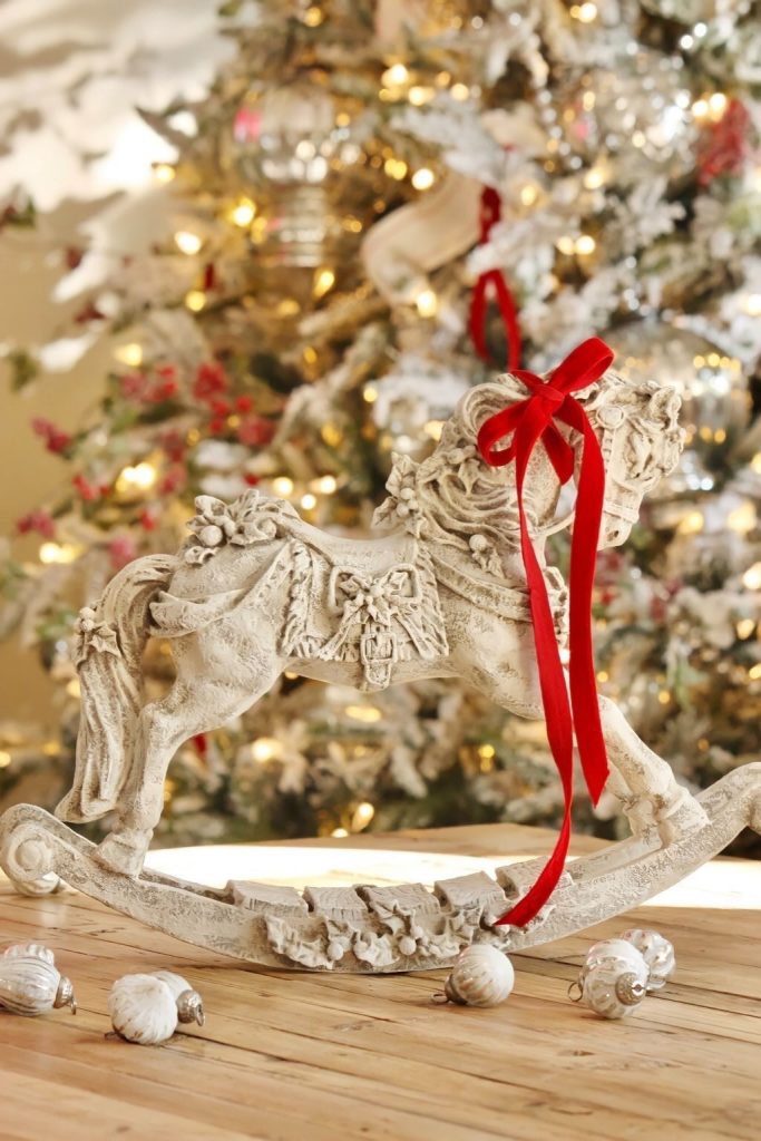 An antiqued rocking horse with red ribbon around the neck sits on a table in front of a Christmas tree.