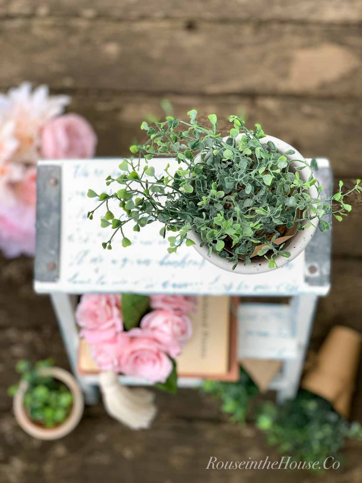 An overhead shot of a step ladder turned into a decorative plant stand. Small faux plants sit on the top and bottom. A small planter of pink peonies sit on the first step.