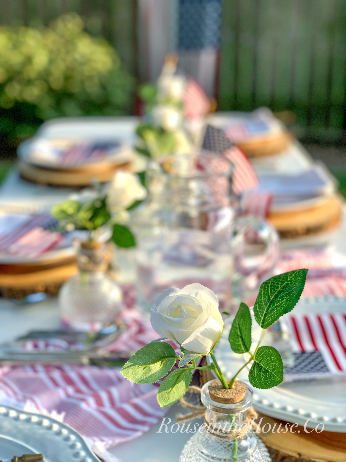 A white rose arrangement is part of a centerpiece on a patriotic tablescape on the patio.  An American flag hangs in the background on the fence.