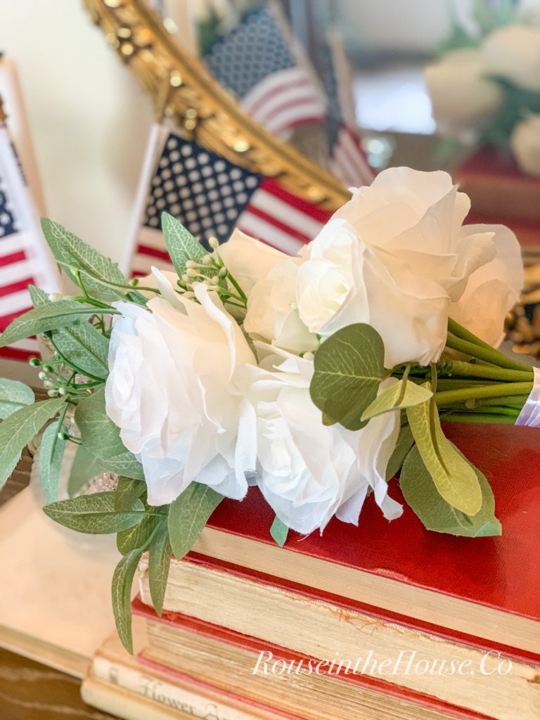 White roses and US Flags grace a stack of red, vintage books near a gold mirror.