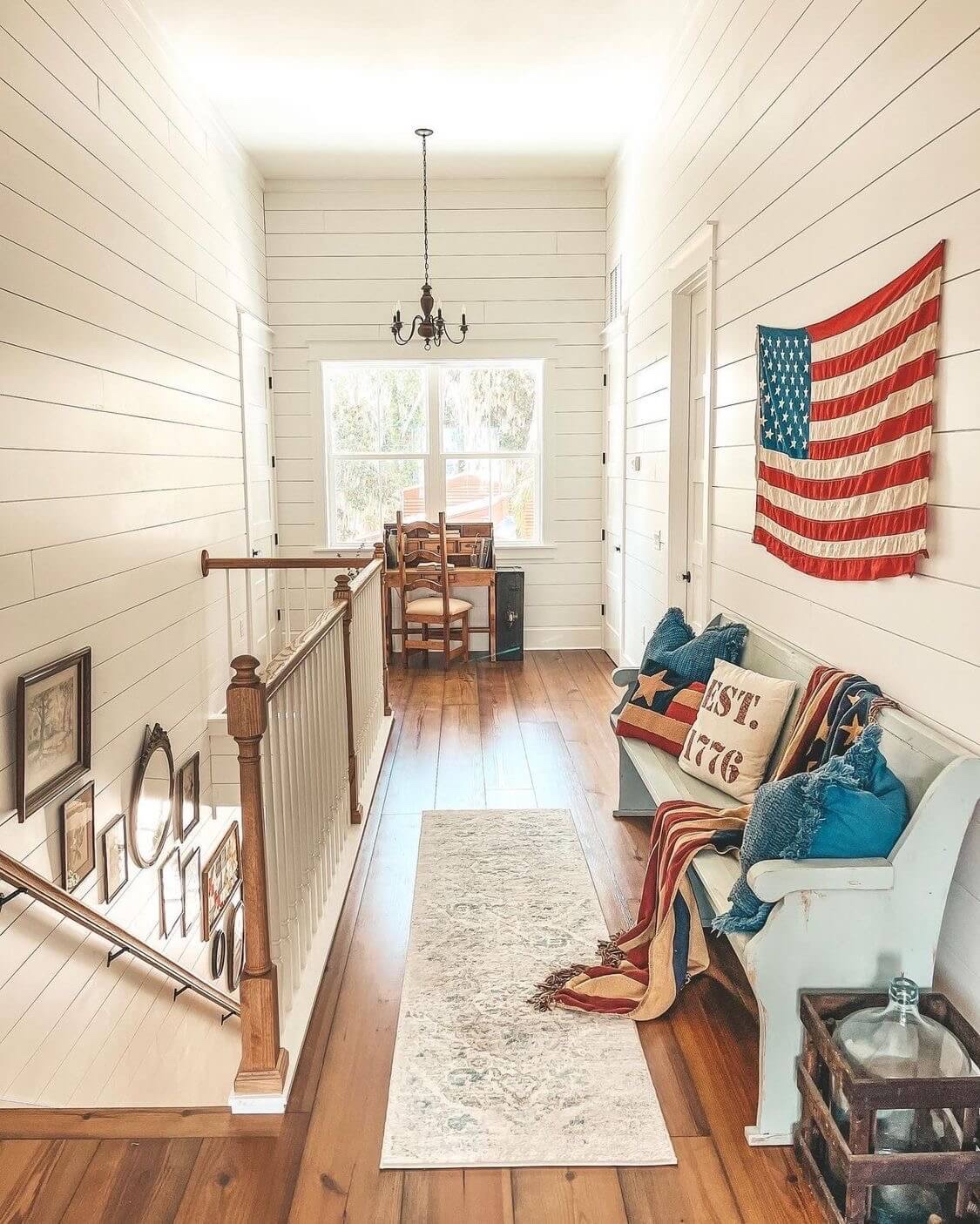 A upper story loft with white shiplap walls and real wood floors is decorated with a large American flag.  Below it, an old church pew is decorated with Americana pillows and a throw.  The adjacent wall is decorated by a wall gallery with antique frames.
