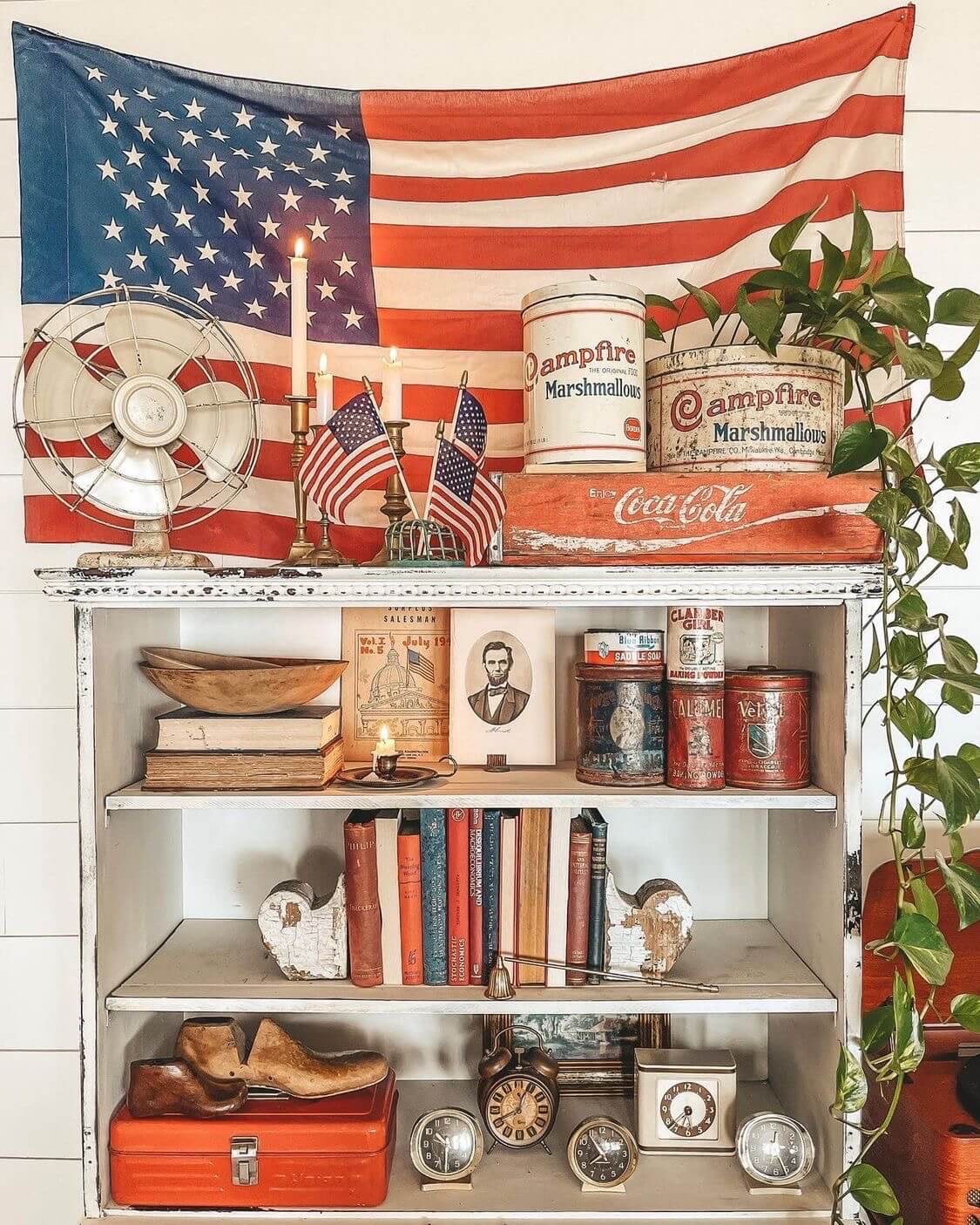 A distressed white shelf is dressed with various antiques including red and blue vintage books, chippy architectural pieces, worn vintage cans, a vintage fan, a black and white portrait of Abraham Lincoln. The top shelf holds a vintage fan, brass candle sticks, small flags, a coca cola wood crate in front a vintage US flag.