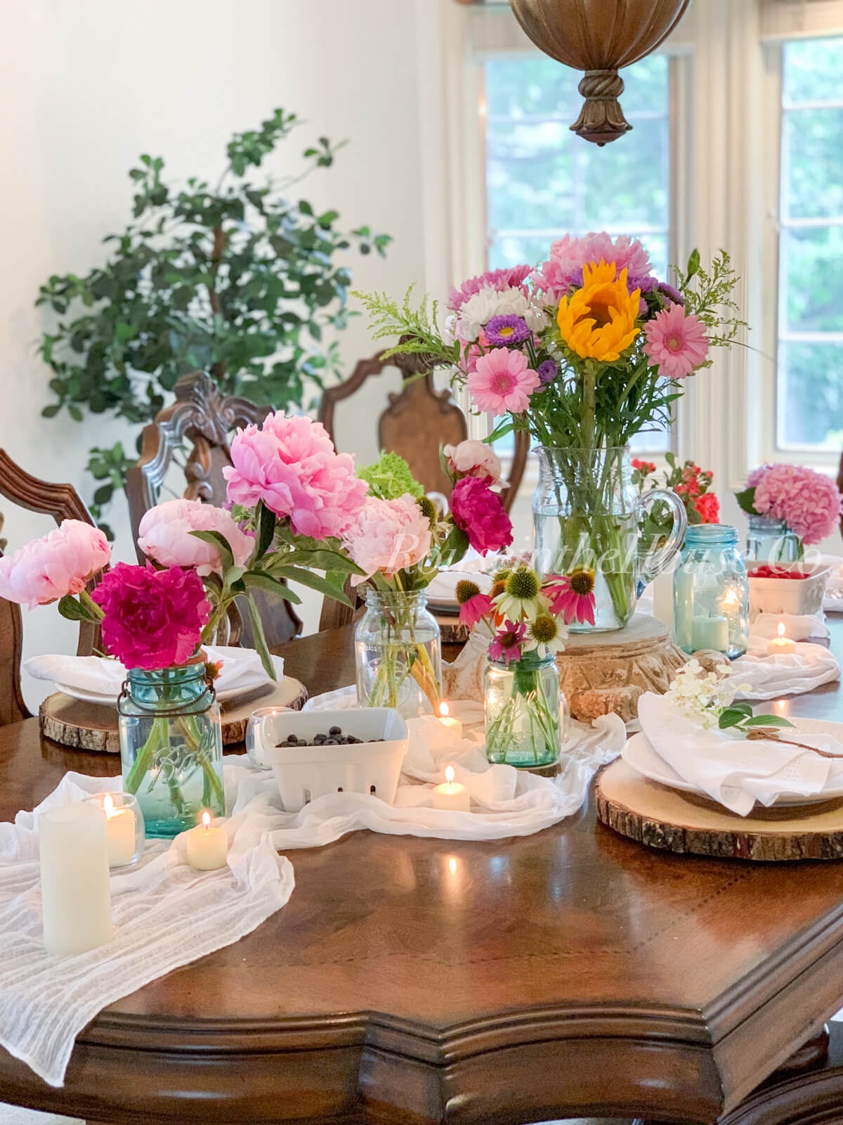 A romantic, country tablescape. A white muslin table runner is draped over a wood table top. Peony, hydrangea, coneflower and mixed flower arrangements in mason jars are along the runner. A large arrangement in a country pitcher is on a distressed pedestal in the center of the table.