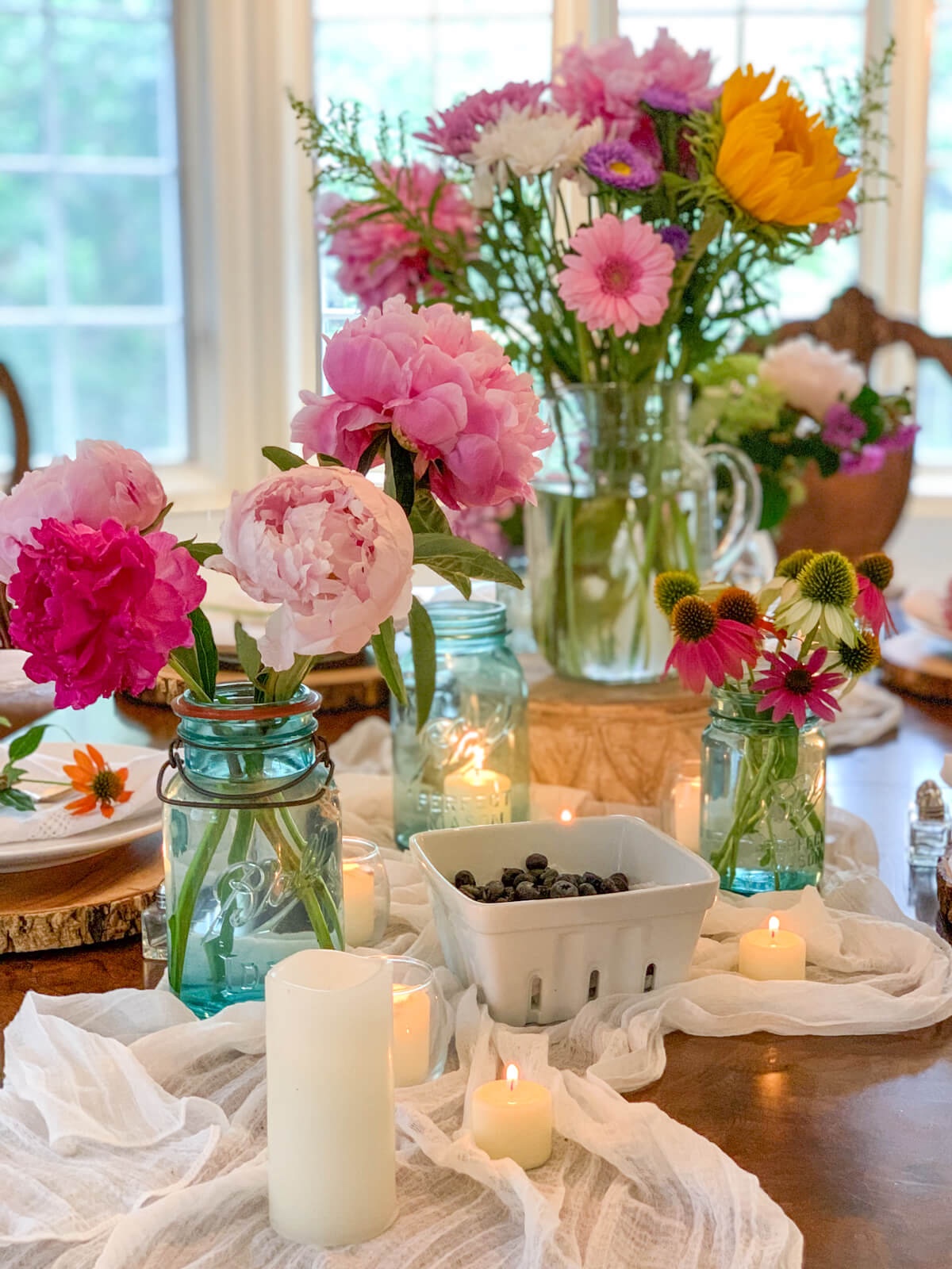 Pink peonies and other flowers are in multiple jars sit next to berries in a berry crate on a romantic tablescape.