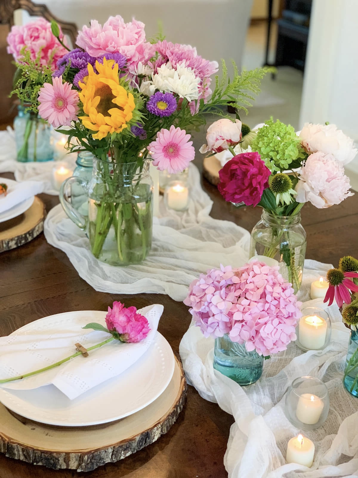 A place setting with a wood charger sits beside a summery country centerpiece with several flower arrangements in mason jars.