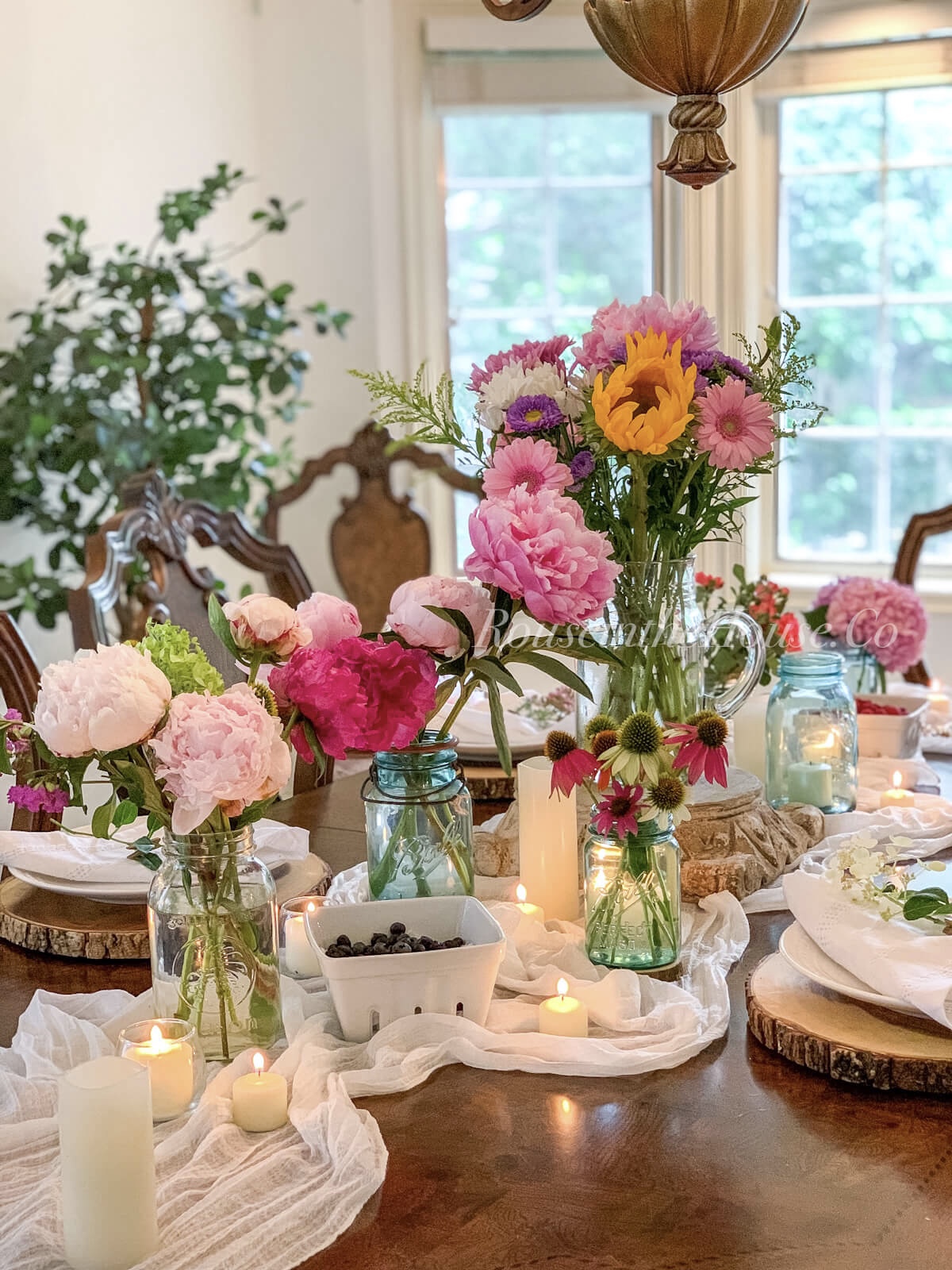 Small & medium arrangements of summer flowers in mason jars sit next to lit votive candles and berries in white berry crates. A beautiful sunflower, daisy and peony arrangement is in the center of the table.