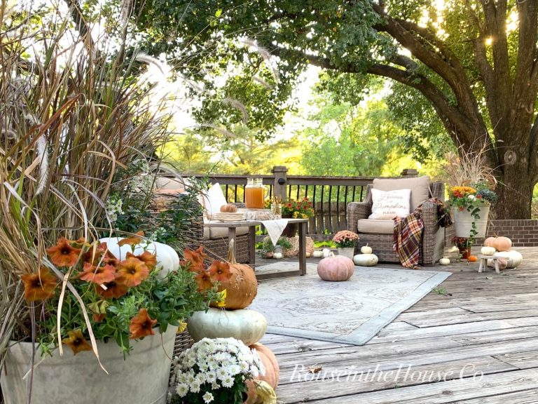 A conversational seating area on a backyard deck is decorated for fall with fall planters, mums, and pretty pumpkins. The outdoor coffee table is styled with a pretty grain sack hand towel, a plate stack, gold flatware and a glass pitcher of apple cider.