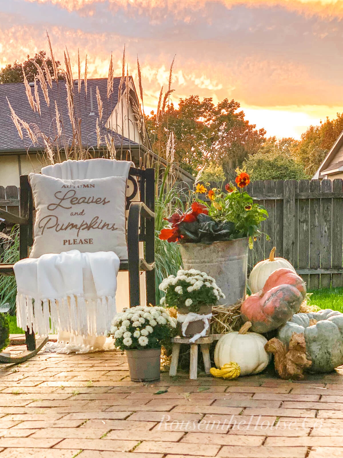 An outdoor fall display on a backyard patio: a square haybale creates a base for an outdoor fall vignette with gourds, pumpkins, small mums in pots and an outdoor fall container.  The outdoor display is next to a rocking chair with a pillow that reads, "Autumn Leaves and Pumpkins Please".