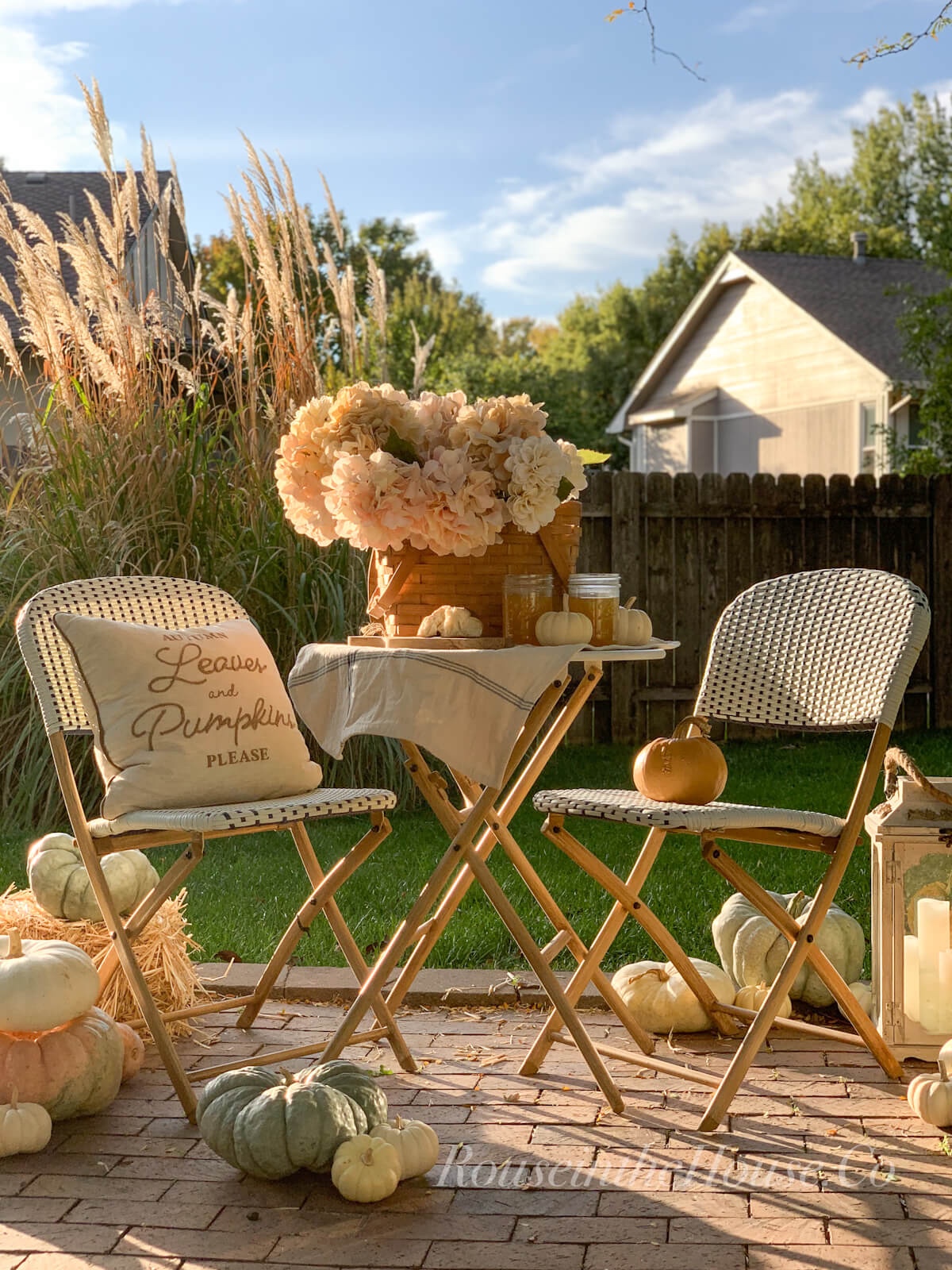 A folding bistro patio set is set up on a brick patio and set with a basket of hydrangeas, apple cider, bread and pretty pumpkins for an outdoor picnic.