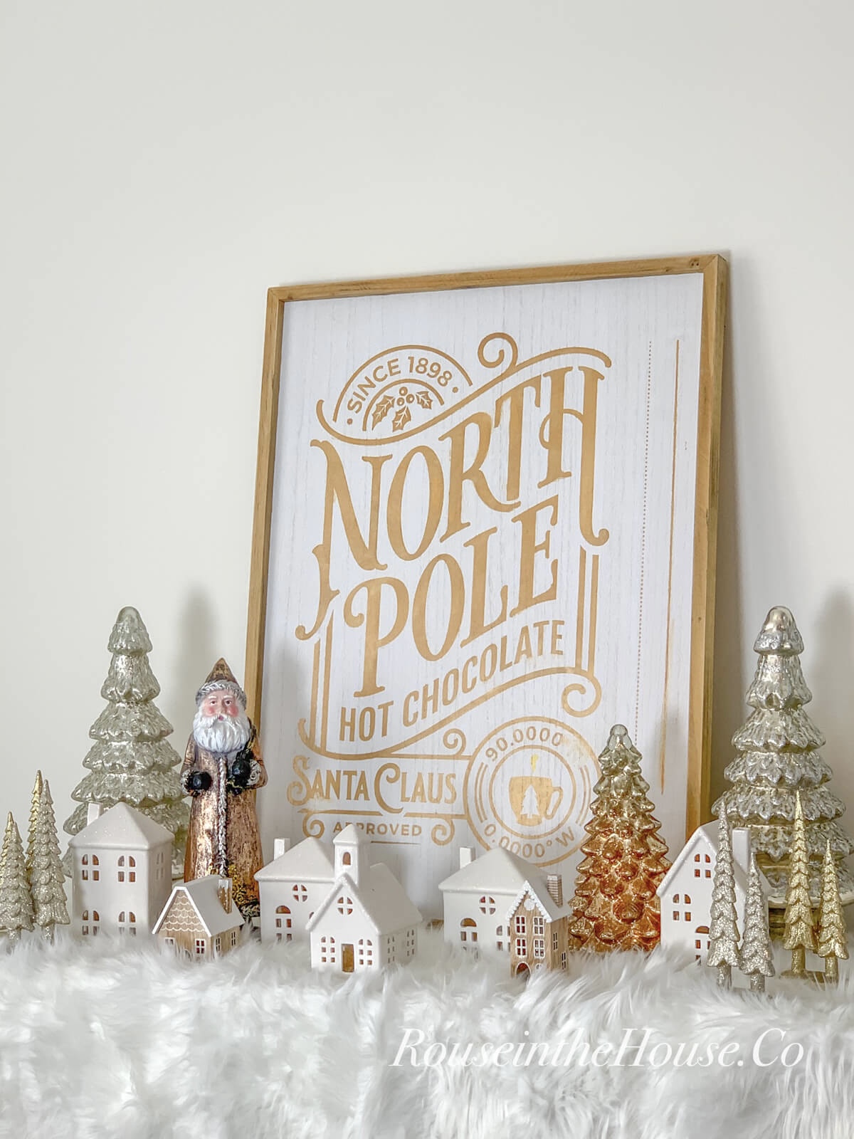 A large Christmas sign that says, "North Pole Hot Chocolate" is sitting on a buffet and surrounded by Christmas village houses, mercury glass trees, and a vintage style Santa.
