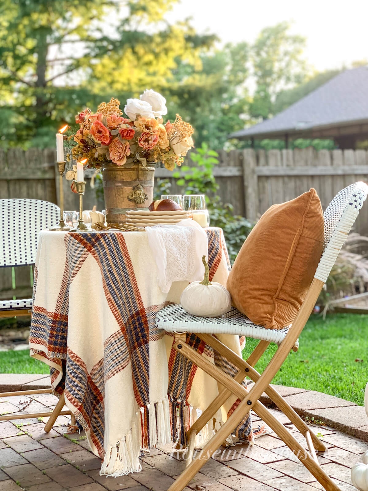 Fall patio decor: Fall Tablescape on Brick Patio. Faux fall flowers in a vintage copper bucket, plates, and brass candlesticks create a fall centerpiece on an outdoor table on the patio.  The woven chair holds a fall corduroy pillow and velvet pumpkin.