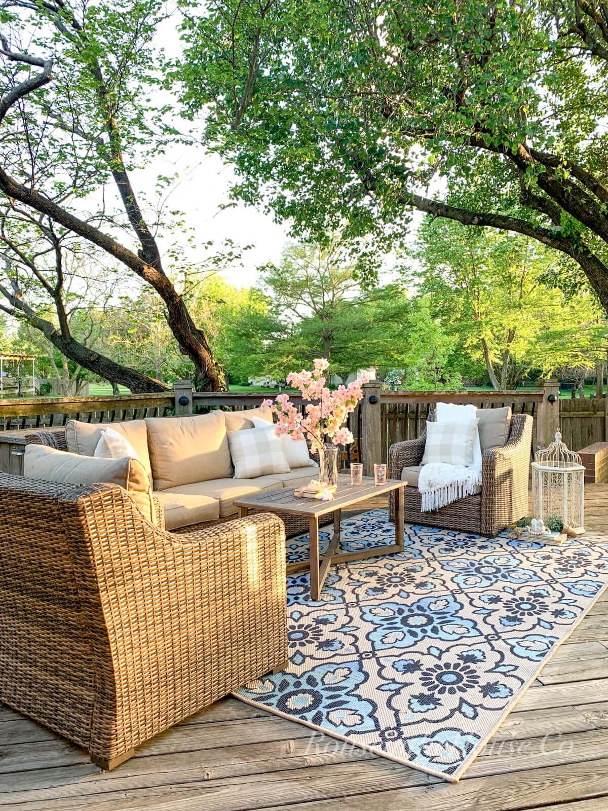 A deck, furnished with outdoor furniture and a large outdoor rug and decorated with throw pillows, a throw blanket and a bird cage as decor.