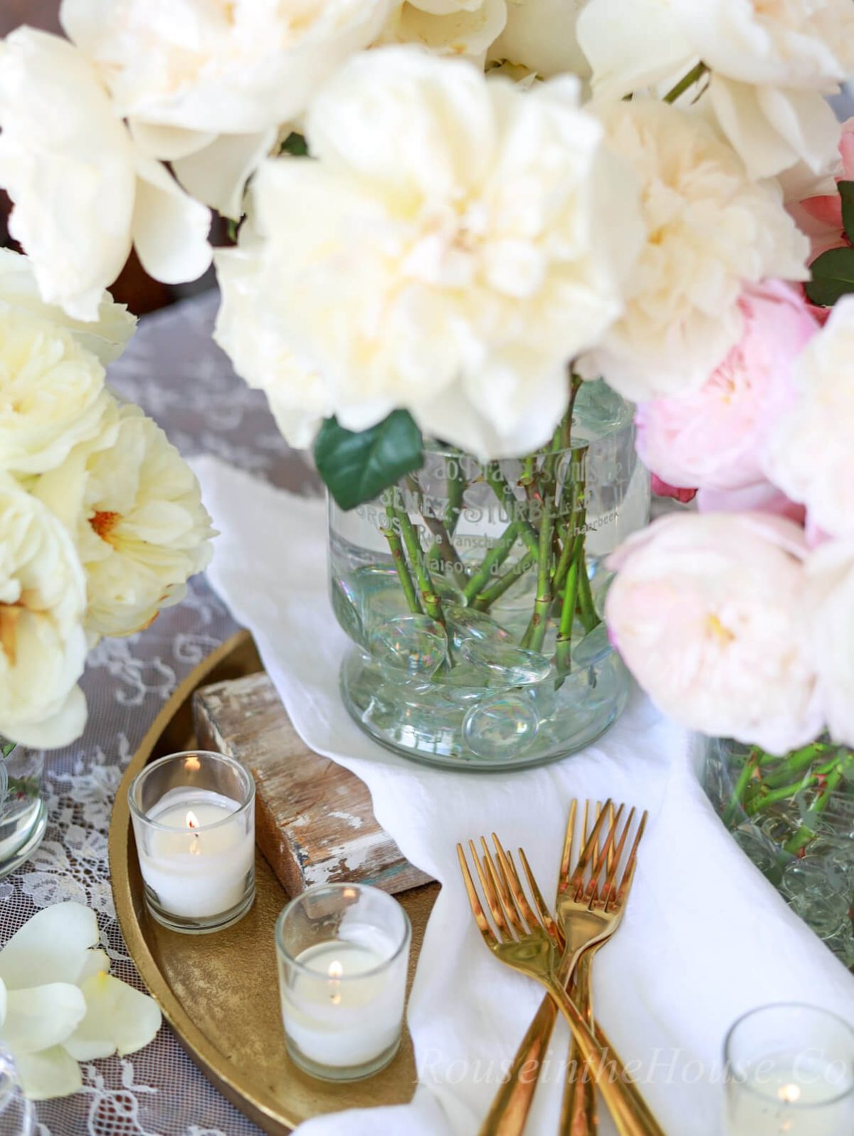 Rose arrangements in etched flower vases are gathered on a gold tray in the center of a dining table along with lit votive candles, gold flatware, and white textiles.
