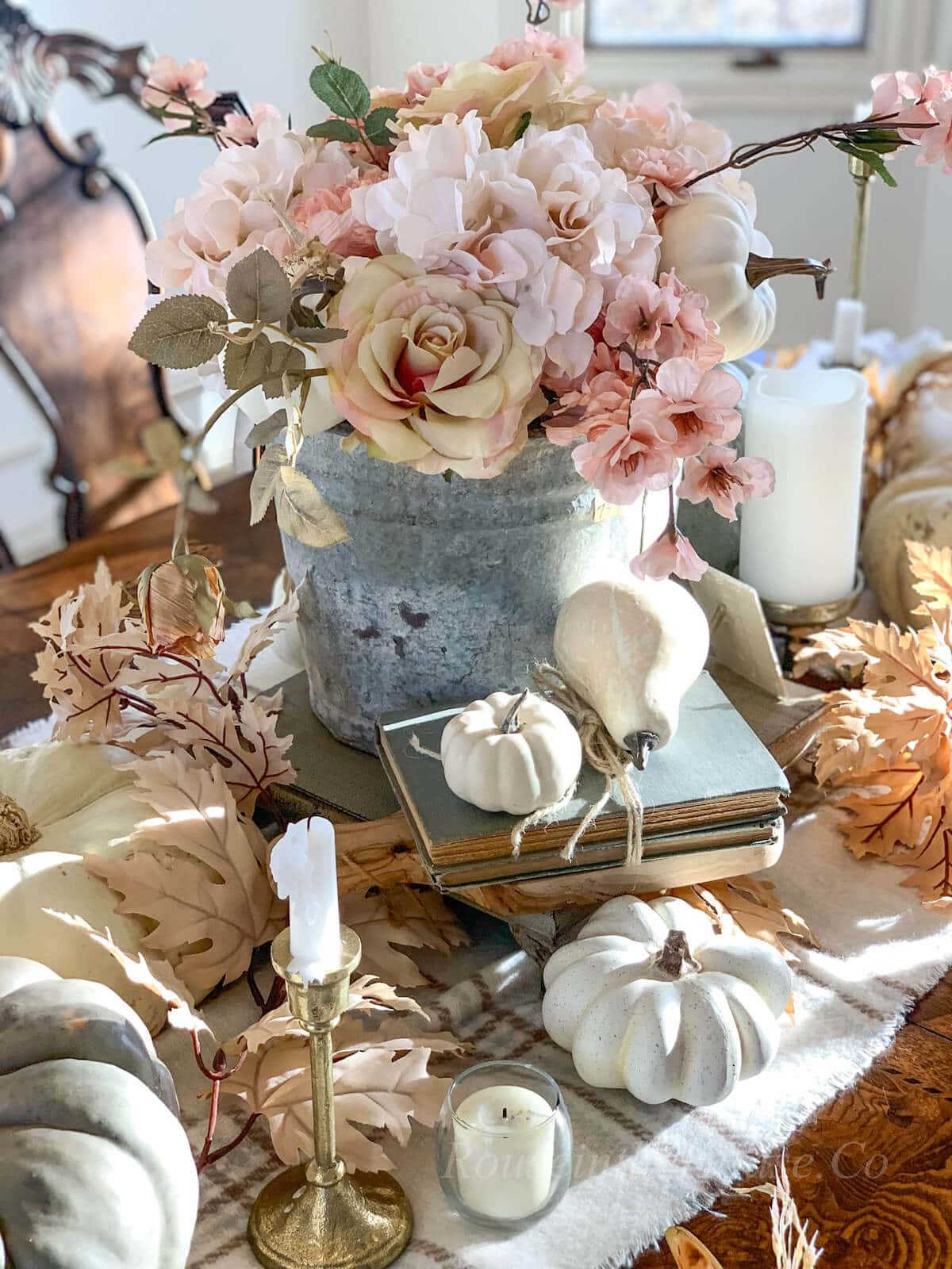 A Thanksgiving centerpiece consists of a fall flower arrangement in a galvanized bucket on a serving tray. A stack of vintage book accents with arrangement. Faux pumpkins and pears are resting on the books and table runner.