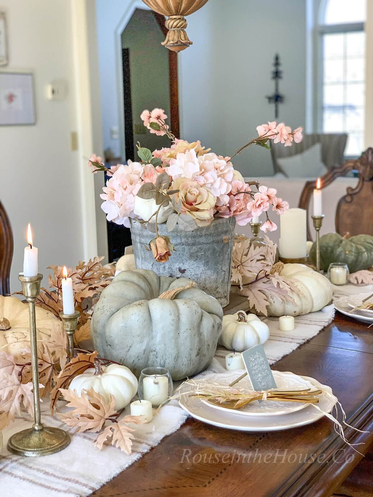 View of a Friendsgiving tablescape: muted pumpkins on a flannel scarf as a table runner, gold candlesticks with lit tapered candles, fall flower arrangement in vintage bucket, and neutral fall leaves tucked in between. 